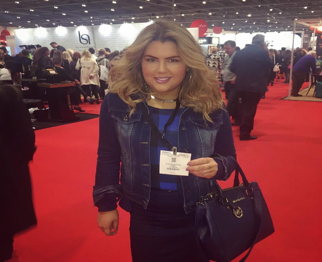 Eine Frau in einem roten Kleid steht auf einem roten Teppich, lacht und hält eine Handtasche, während Fotografen und Scheinwerfer im Hintergrund zu sehen sind.