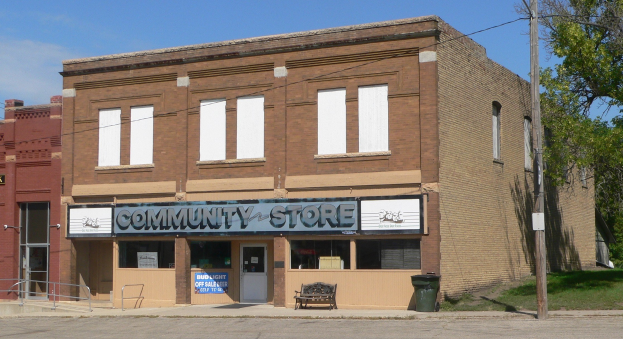 Ein einstöckiges Gebäude mit der Aufschrift "Community Store", einer Bank, einem Mülleimer und umgebender Vegetation unter einem bewölkten Himmel.