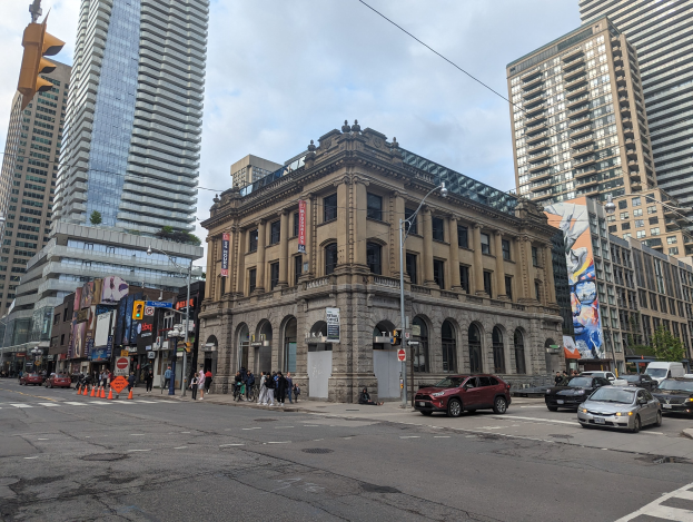 Eine belebte Stadtstraße mit hohen Gebäuden, Fahrzeugen, Fußgängern, Verkehrsinfrastruktur und der Toronto Public Library als Mittelpunkt unter einem bewölkten Himmel.