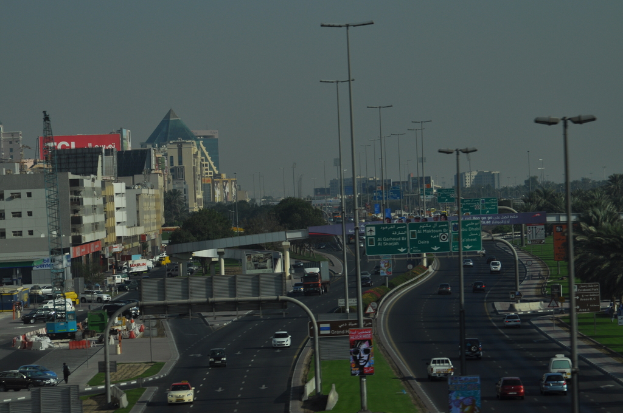 Eine Luftaufnahme einer Stadt mit Gebäuden, Türmen, Fahrzeugen, Werbetafeln, Schildern, Straßen, Bäumen, Straßenlaternen, Menschen, Verkehrskegeln und Informationsanzeigen unter einem klaren Himmel.
