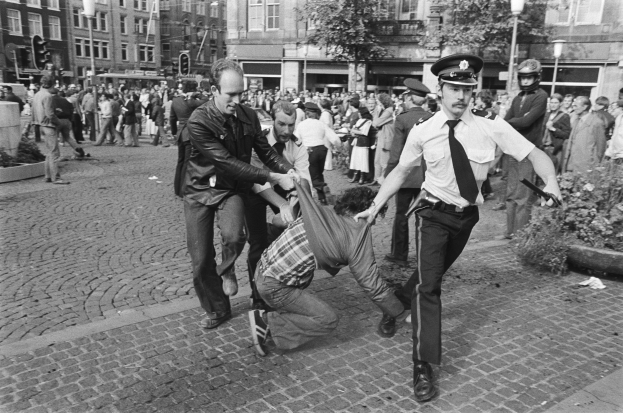 Schwarz-weißes Bild eines Mannes, der von zwei Polizisten auf einer Straße mit Zuschauern, Bäumen, Gebäuden, Laternen, Verkehrsampeln, Topfpflanzen und einem Mann im Vordergrund mit Schuhen gezogen wird.