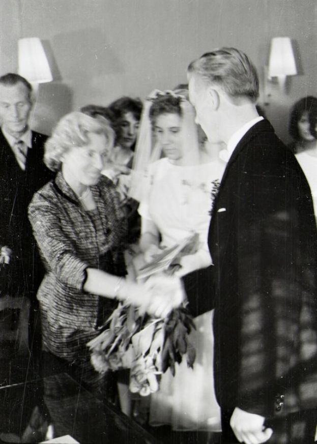 Ein Schwarz-Weiß-Foto eines Brautpaares, das sich die Eheringe reicht, mit der Braut in einem weißen Kleid und Schleier, die einen Blumenstrauß hält, und dem Bräutigam in einem Anzug und Krawatte, mit Wandlampen im Hintergrund.