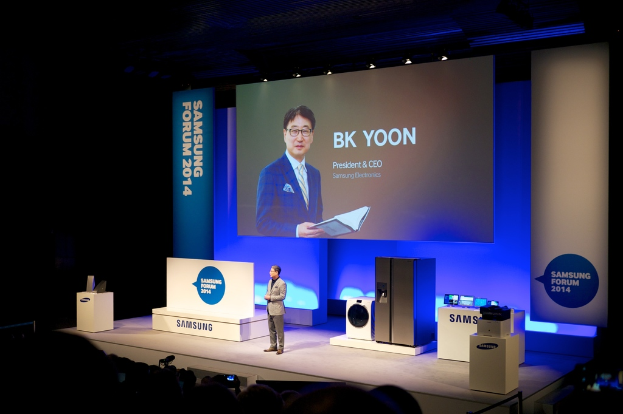 Ein Mann steht vor einer Tafel und einem kühlschrankähnlichen Objekt, hält ein Buch und lächelt, während Menschen auf Stühlen in einem Konzertsaal mit beleuchteter Decke sitzen.
