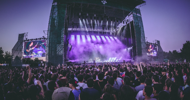 Eine große Menschenmenge auf einem Musikfestival schaut Künstlern auf einer hell erleuchteten Bühne mit Instrumenten und Bildschirmen zu, die vor einem Gebäude, Bäumen und einem klaren blauen Himmel liegt.