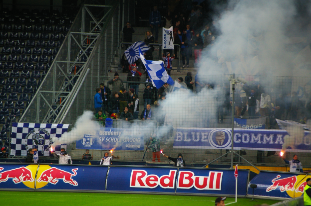 Eine Gruppe von Menschen steht auf einem Fußballfeld und hält Fahnen und Schilder hoch, umgeben von einem Metallzaun mit Rauch, der aufsteigt, und leeren Stühlen auf der linken Seite.