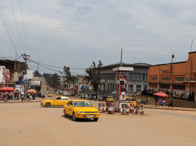 Ein gelbes Taxi fährt auf einer belebten Stadtstraße mit Gebäuden an den Seiten, während Fußgänger mit Schirmen auf dem Gehweg darunter gehen, bei bewölktem Himmel, mit Strommasten und Leitungen im Hintergrund.