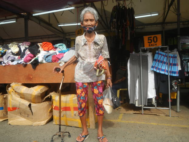 Eine ältere Frau mit Gesichtsmaske steht an einem Marktstand und hält einen Gehstock und eine Decke in der Hand, während hinter ihr Kleidung auf einem Tisch und an nummerierten Bügeln hängt, und im Hintergrund sitzt eine Person unter Deckenleuchten.