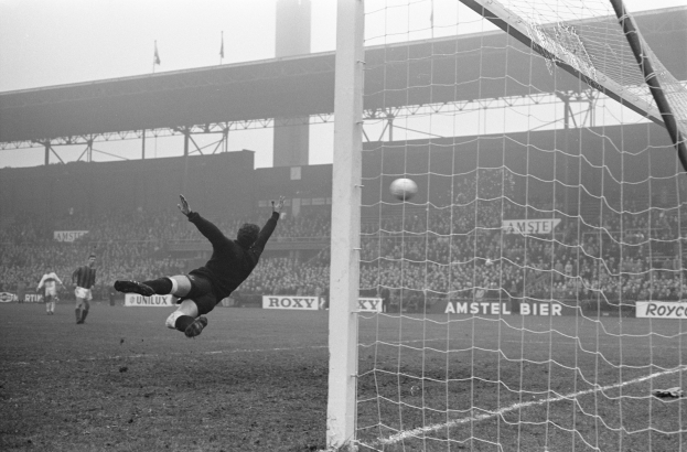 Ein Schwarz-Weiß-Foto eines Fußballspielers, der vor einem Tor einen Abschlag macht, mit einem Netz auf der rechten Seite und einer Zuschauermenge im Hintergrund, sowie Schildern mit Text, einer Brücke und Fahnen mit Stangen.