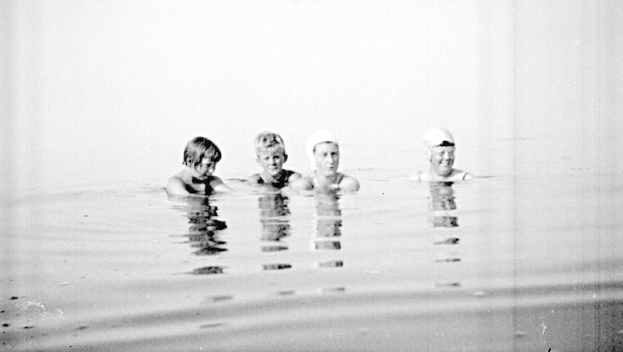 Schwarz-weiß-Foto von vier Kindern, die in einem Pool schwimmen, alle tragen Schwimmkappen.