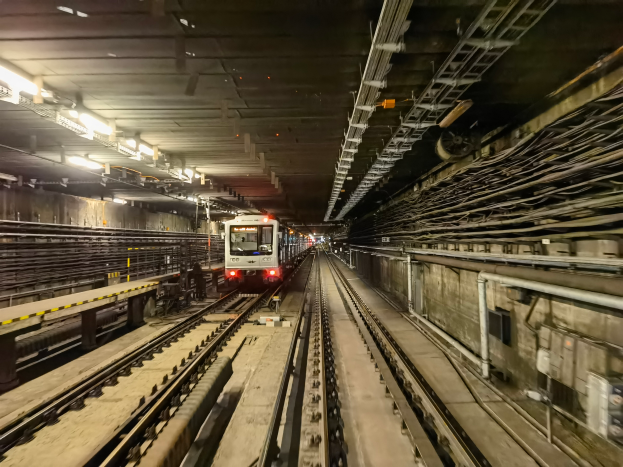 Ein Zug fährt durch eine hell erleuchtete U-Bahn-Station in der Nacht, mit Plattformen, Gleisen und Kabeln auf beiden Seiten.