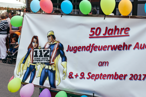 Eine Gruppe von Menschen auf einer Straße, einige halten Kinderwagen, mit einem Banner "50 Jahre Jugendfeuerwehr August 2017" mit Text und Cartoon-Bildern, einem Zaun, Ballons, einem Zelt, einem Gebäude mit Fenstern und Bäumen im Hintergrund.