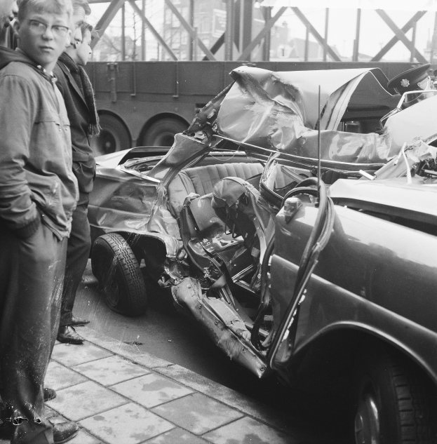 Schwarz-weißes Bild eines verunglückten Autos am Straßenrand mit mehreren Menschen in der Nähe, einem Lastwagen und einer Brücke im Hintergrund.