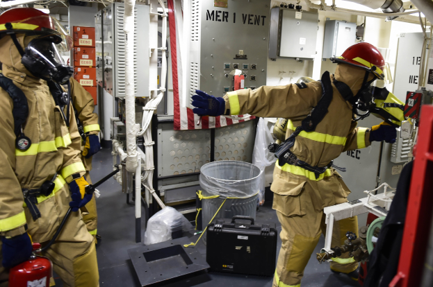 Drei Feuerwehrleute in Schutzausrüstung stehen zusammen in einem Raum mit einem Feuerlöscher links, einer Maschine rechts und verschiedenen Gegenständen auf dem Boden.