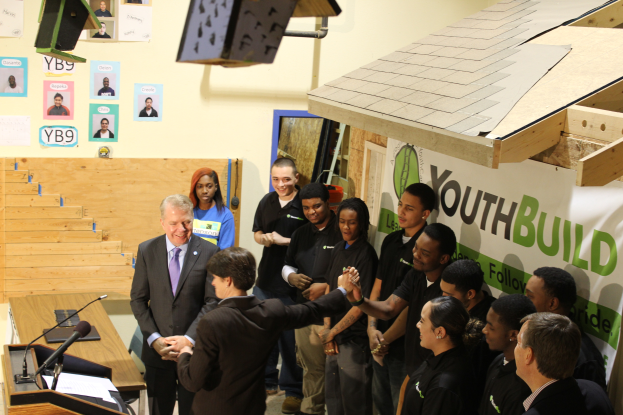 Eine Gruppe von Menschen steht um ein Rednerpult herum vor einer Menge, einige schütteln Hände, ein Tisch mit Mikrofonen und Papieren links, Plakate an der Wand im Hintergrund und Gegenstände auf dem Dach und der rechten Seite.