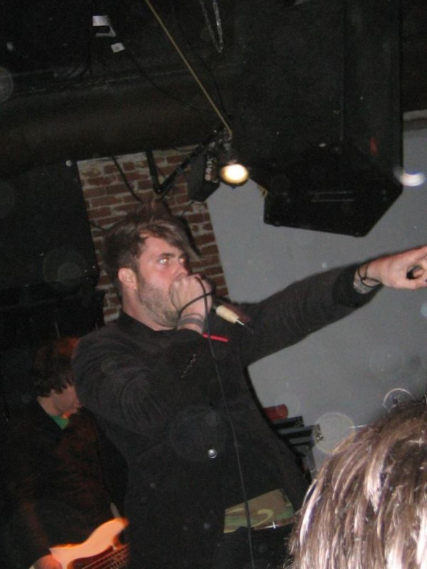 Ein Mann singt in ein Mikrofon in der Mitte einer Gruppe von Menschen, mit Lichtern im Hintergrund.