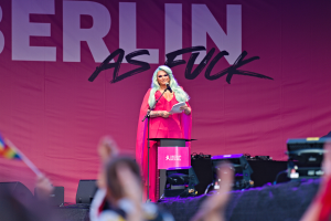 Eine Frau in einem rosa Kleid steht an einem Podium mit einem Mikrofon und Papier und spricht zu einer Menge mit einem "Berlin as Fuck"-Plakat im Hintergrund.