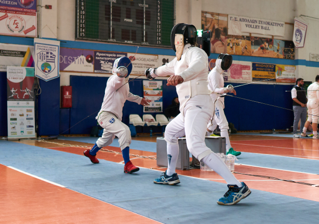 Eine Gruppe von Menschen in Helmen und mit Schwertern ficht in einer Turnhalle mit Bannern, einem Bildschirm, Stühlen und anderen Gegenständen im Hintergrund.
