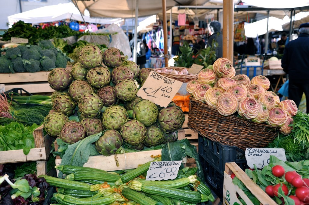 Körbe voller Obst, Gemüse und kleine Schilder mit Text, mit Zelten und ein paar Menschen im Hintergrund.