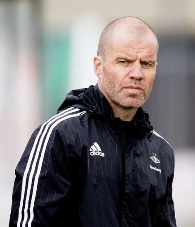 Glatzköpfiger Trainer in marineblauer Jacke mit weißen Streifen und weißem Kragen auf einem Fußballfeld stehend, Hände verschränkt, ernster Gesichtsausdruck, unscharfer Hintergrund.