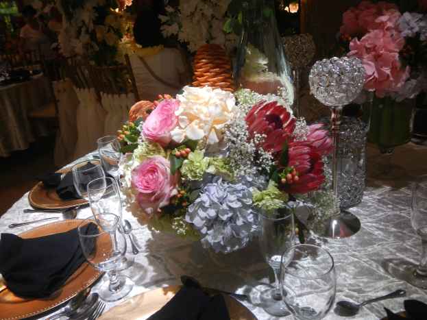 Ein formeller Esstisch mit Tellern, Besteck, Gläsern, Servietten und Blumenvasen, mit einem floralen Zentrum und Stühlen und Tischen im Hintergrund.