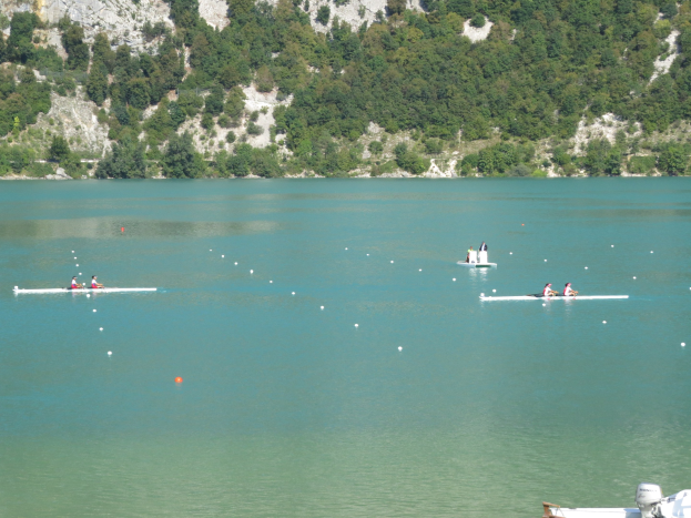 Gruppe von Menschen, die in einem Rennen auf einem Gewässer mit einem von Bäumen bedeckten Hügel im Hintergrund rudern.