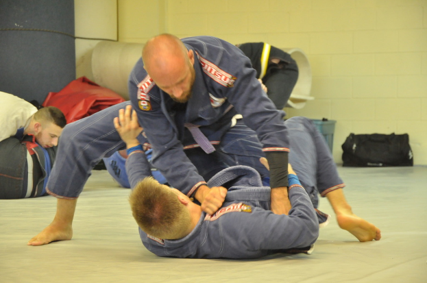 Zwei Personen, die Kampfsport betreiben, während zwei weitere auf der linken Seite zuschauen, ein Sack auf der rechten Seite und eine Wand im Hintergrund.
