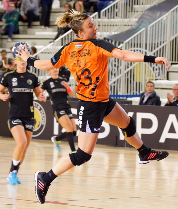 Eine Frau in einem orangen T-Shirt und schwarzen Shorts hält einen Handball, mit Zuschauern auf Treppen und Geländern im Hintergrund und einem Banner mit der Aufschrift "Krp Slezak - Handball-Weltmeisterschaft 2019."