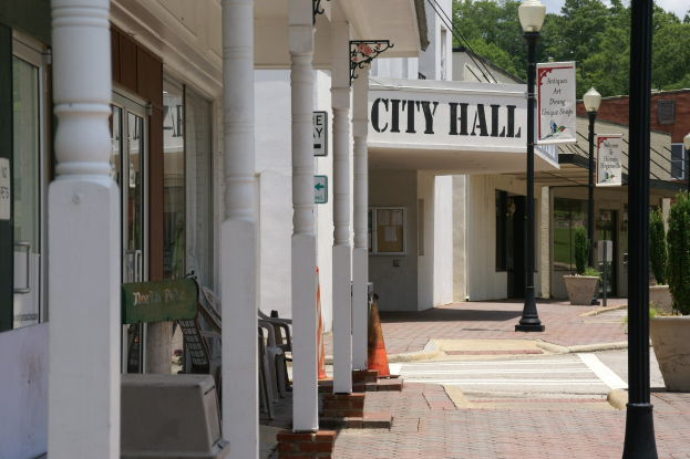 Rathausgebäude mit umliegender Infrastruktur, einschließlich Straßenlaternen, Schildern, Fahrzeugen, Absperrgittern, Stühlen, Topfpflanzen, Bäumen und einer bewölkten Himmel.