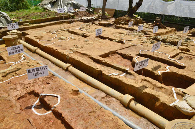 Großes Erdaushubgelände mit Erde, umgeben von Bäumen, Pflanzen, Gras, einem Zaun, einer Abdeckung und einer Wand, mit Schildern, Rohren und verstreuten Gegenständen.