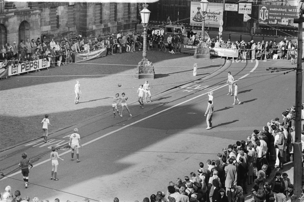 Schwarzes Bild eines Streetsoccer-Spiels mit Spielern im Einsatz und Zuschauern von der Seitenlinie aus, umgeben von Laternenmasten, Gebäuden und Bannern.