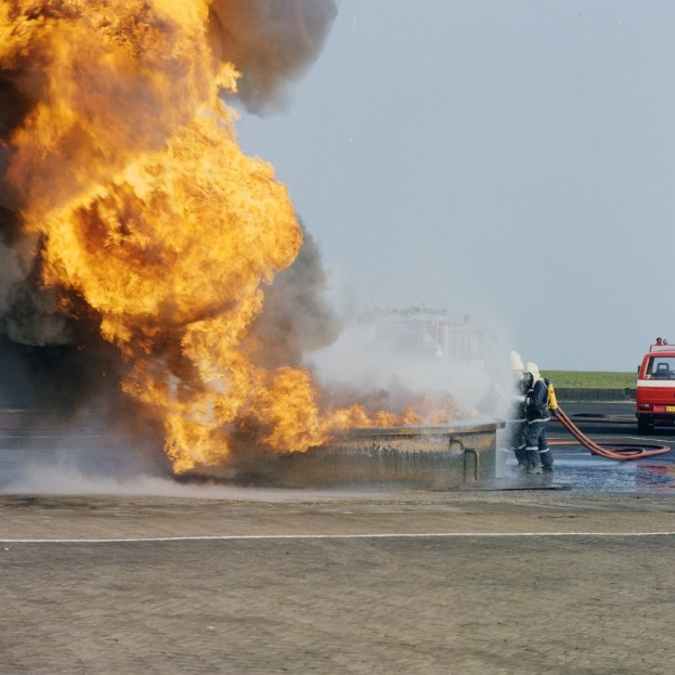 Feuerwehrlöschfahrzeug von Flammen umgeben auf der Straßenseite mit zwei Personen in Helmen und Schläuchen, einem Fahrzeug im Hintergrund und dem Himmel.