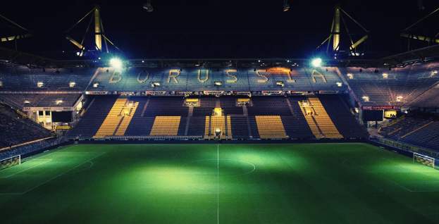 Ein Fußballfeld bei Nacht, beleuchtet von hellen Stadionlichtern, mit saftig grünem Rasen und einigen Menschen auf den Tribünen.