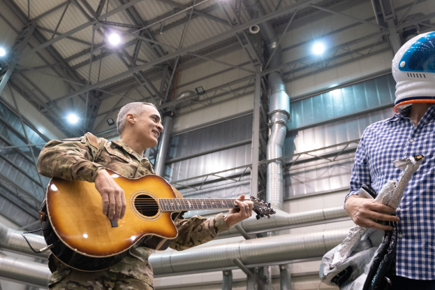 Ein Mann in militärischer Uniform spielt eine akustische Gitarre, während er neben einem anderen Mann in einem Helm steht, mit industriellen Rohren und Deckenlampen im Hintergrund.