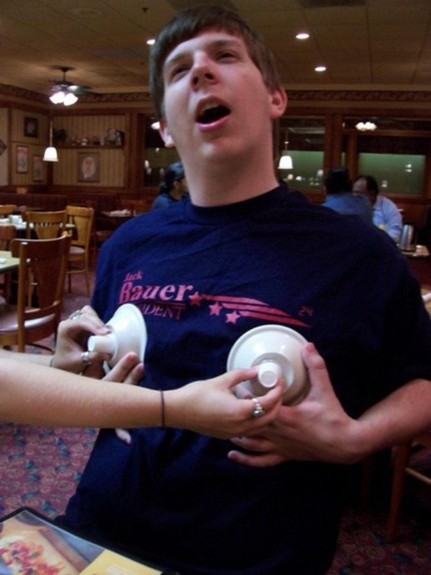 Ein Mann in einem blauen Hemd hält Tassen in einem Restaurant, mit zwei Personen an einem Tisch und Lampen an der Decke im Hintergrund.