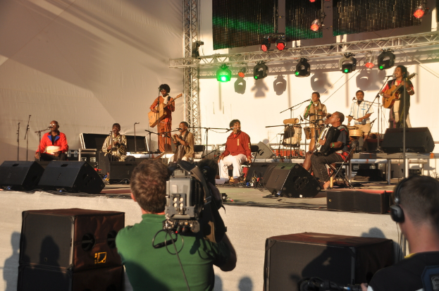 Ein musikalisches Konzert mit Musikern, die verschiedene Instrumente wie Gitarren und Trommeln spielen, auf der Bühne unter rotem und grünem Scheinwerferlicht, mit Lautsprechern unten und zwei Personen, eine Aufnahme und die andere zuhört.