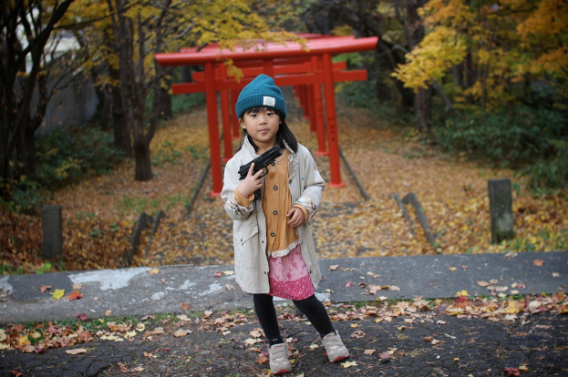 Ein Mädchen steht auf einer Straße und hält eine Waffe in der Hand, trägt eine graue Jacke und hat Bäume und rote Metallobjekte im Hintergrund sowie trockene Blätter auf dem Boden.
