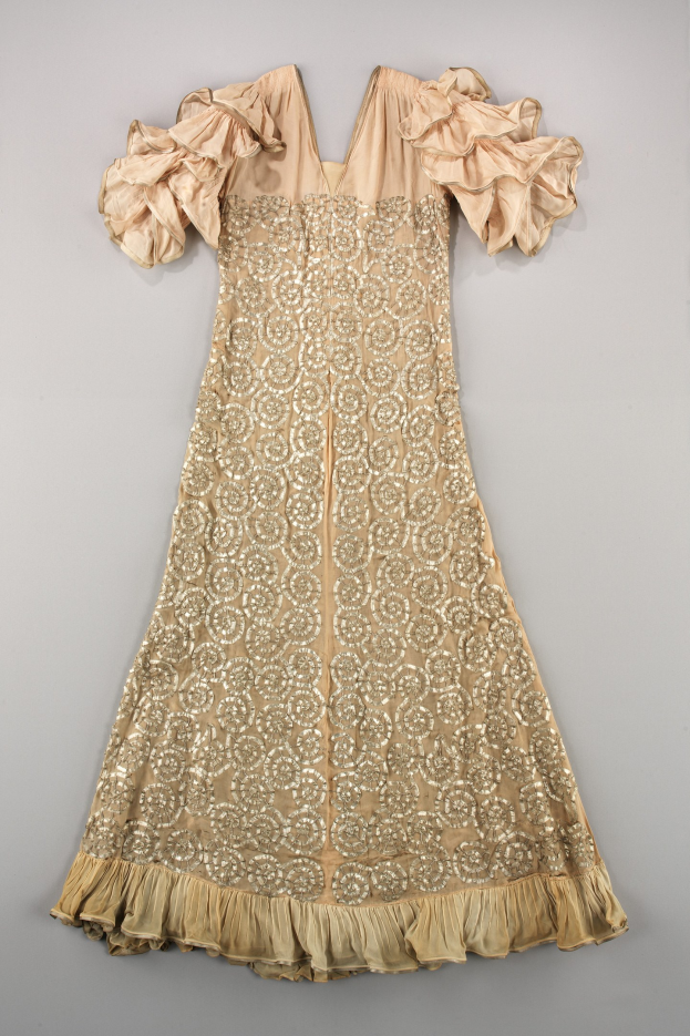 Ein cremefarbenes Kleid mit goldener Stickerei und Rüschenärmeln, das auf einer weißen Oberfläche präsentiert wird und detaillierte Muster und Designs aufweist.