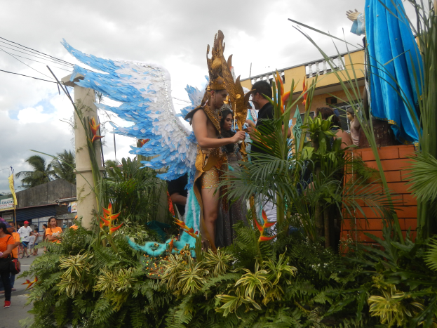 Eine Gruppe von Menschen geht neben einem Karnevalswagen mit Pflanzen und Blumen, kostümierten Personen auf dem Wagen, Gebäuden, Bäumen und Strommasten mit Drähten im Hintergrund und einem bewölkten Himmel.