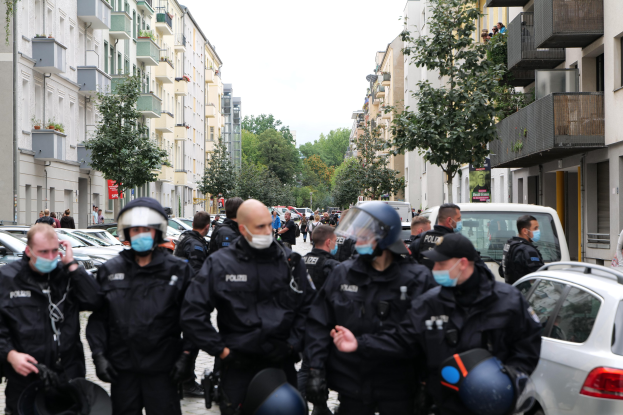 Eine Gruppe von Polizisten in Masken und Helmen geht eine Straße mit geparkten Autos entlang, flankiert von Gebäuden mit Balkonen und Geländern, unter einem klaren Himmel mit Bäumen im Hintergrund.