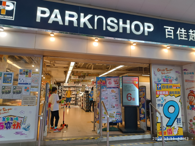 Ein Park-Shop in einem Einkaufszentrum mit einem Schild, auf dem "Park Shop" steht, Menschen auf dem Boden, Metallstangen, Schilder, Banner, Regale mit Gegenständen und Deckenleuchten.