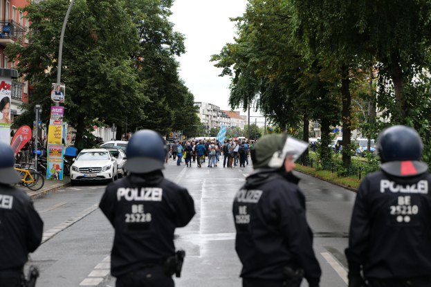 Eine Gruppe von Polizisten in schwarzen Uniformen und Helmen steht auf der Straße, mit Menschen, Fahrzeugen, Fahrrädern, Pfählen, Plakaten, Bäumen, Gebäuden und einem Banner im Hintergrund.