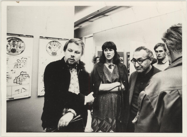 Eine Gruppe von Menschen, die in einem Raum mit einer plakatgeschmückten Wand stehen und sprechen, in Schwarz-Weiß, aus den 1980er Jahren.