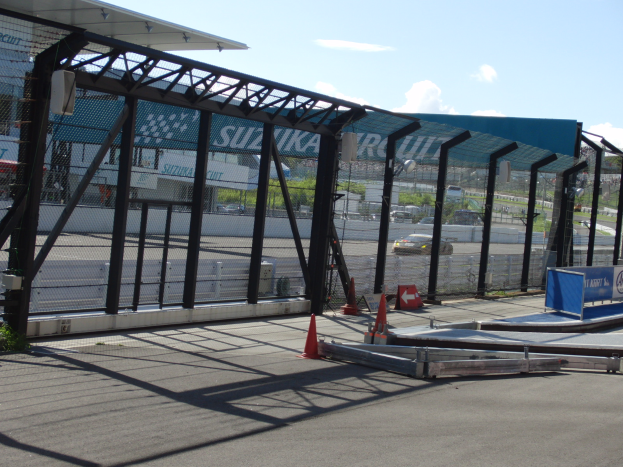 Eine Rennstrecke mit parkenden Autos, Verkehrskegeln, einem Zaun, einem Schuppen, einer Fahne, Pflanzen, Bäumen und einem bewölkten Himmel.