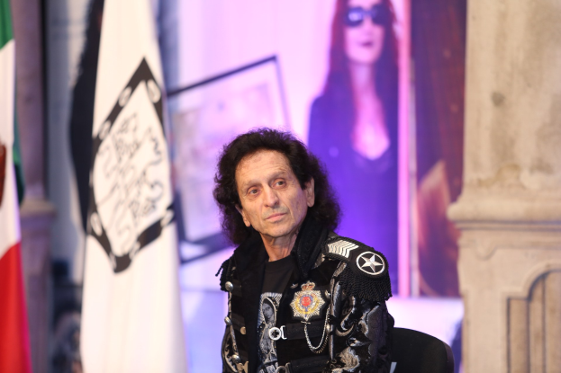 Gene Simmons sitzt in einem Stuhl und schaut in die Kamera, während er von Flaggen umgeben ist.