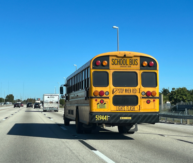 Ein gelber Schulbus fährt auf einer Autobahn mit anderen Fahrzeugen, begrenzt von einem Geländer auf der rechten Seite, Bäumen und Masten im Hintergrund und einem klaren blauen Himmel.