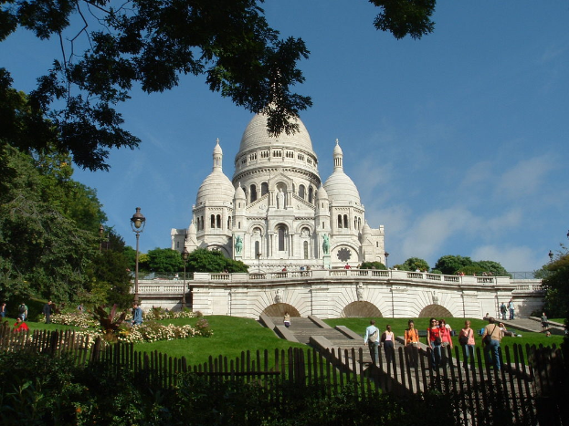 Ein weißes Gebäude mit Menschen, Pflanzen, Zäunen, Gras, Pfählen und Bäumen in der Nähe.