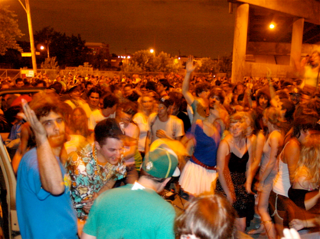 Menschen tanzen im Vordergrund auf einem Konzert, mit Lichtern, Pfählen, Bäumen und anderen Objekten im Hintergrund.