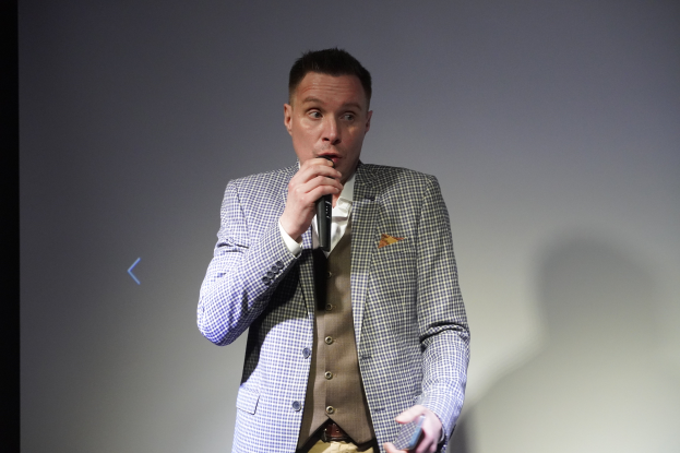 Ein Mann im Anzug steht selbstbewusst auf einer Bühne, hält ein Mikrofon und hat einen entschlossenen Gesichtsausdruck sowie ordentlich gekämmtes Haar.