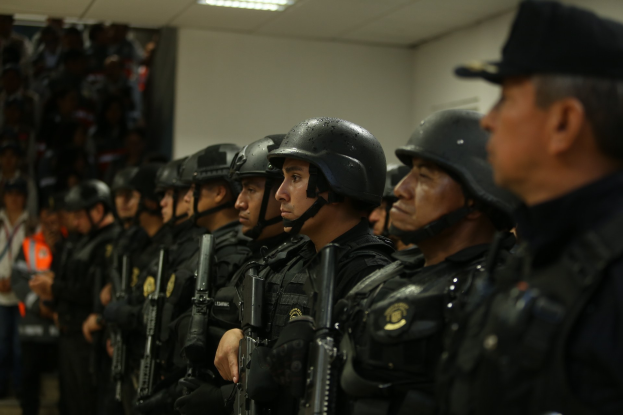 Gruppe von Polizisten in Uniform, die in Formation stehen und Helme sowie Feuerwaffen tragen, vor einer Wand und einer Deckenbeleuchtung.