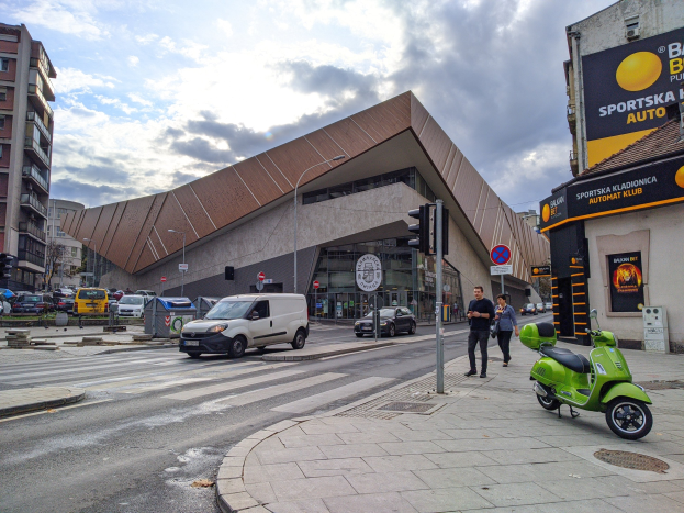 Ein grüner E-Scooter steht auf dem Seitenstreifen neben einem Gebäude, mit Fahrzeugen auf der Straße, Menschen auf dem Gehweg und Ampelmasten mit Verkehrssignalen, mit Gebäuden, Bäumen und einem bewölkten Himmel im Hintergrund.